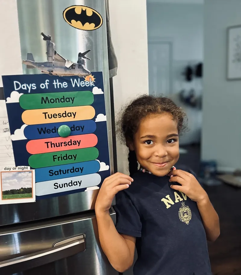 Student with a printed Days of the Week chart held on a refrigerator with magnets