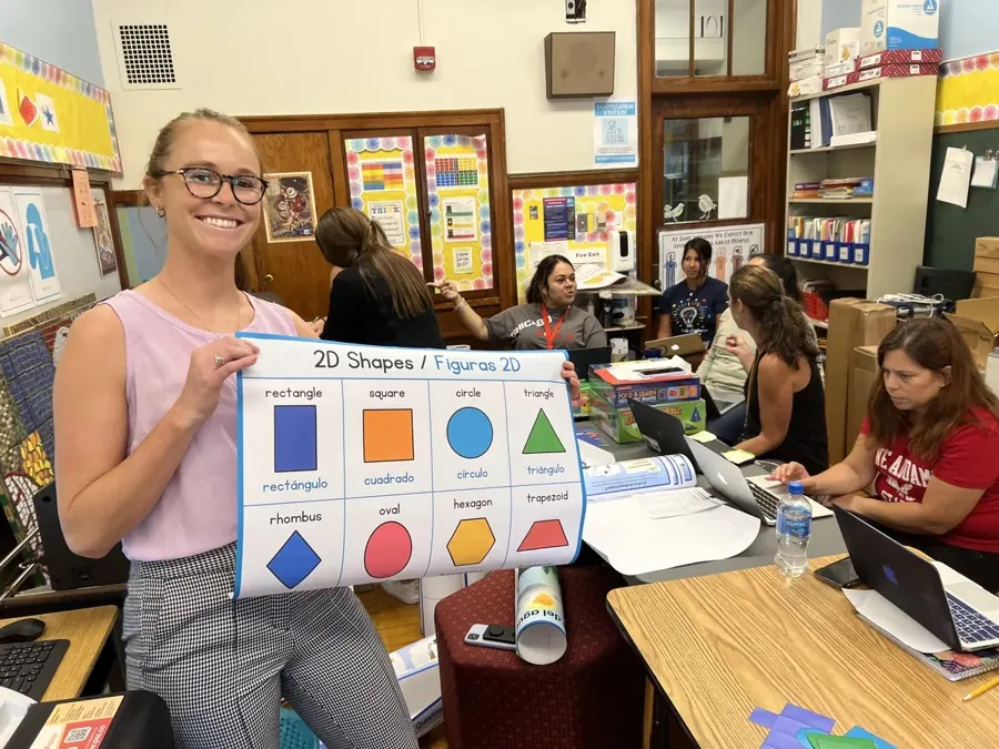 Teacher holding a 2D Shapes poster in English and Spanish printed minutes earlier in her classroom